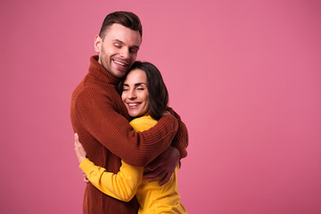 Young cheerful loving couple dressed in sweaters hugging isolated over pink background