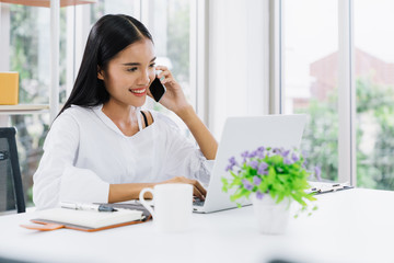 Young asian casual businesswoman using mobile phone and laptop to communicate with customer for order purchasing abd delivering information in modern home office, online shopping business concept