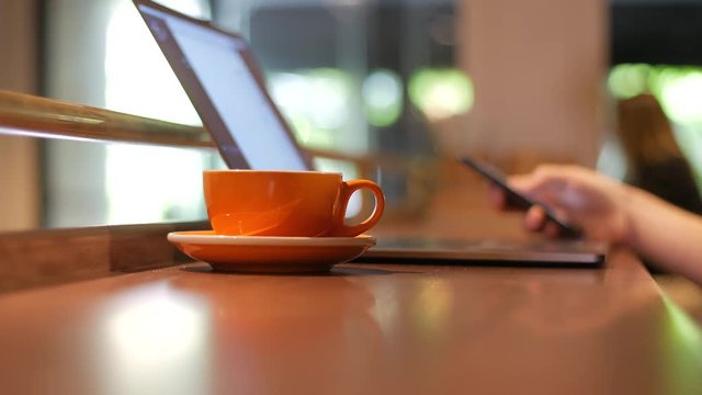 View of coffee cup, laptop and blurred male hand using smartphone at wooden table in coffee shop
