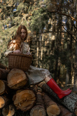 Young woman with long red hair in a linen dress gathering mushrooms in the forest in the bast basket