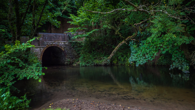 Old dark stone bridge across the river Frome in Maiden Newton, Dorset, UK. Medieval bridge with reflection in the river among green trees. Fairytale place in England.