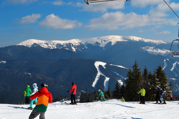 Landscape of winter hills in sunny day. Sport concept. People in colorful sportwear are skiing down the slope under below chairlift with skiers and snowboarders lifting to top of mountain in Ukraine.