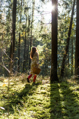 Young woman with long red hair in a linen dress gathering mushrooms in the forest in the bast basket