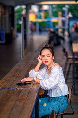 portrait asian girl wearing white shirt and jeans in outdoor sit on the chair in coffee outdoor by holding phone for waiting messages