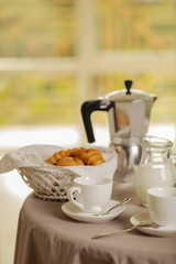 Coffee maker, cups and croissants in the basket are on a round table in front of the window.