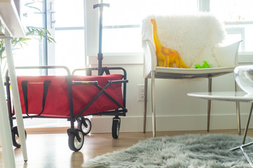 Red foldable wagon in bright apartment living room, with dinosaur toys sitting on a chair. Grey...