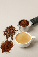 Cup of black coffee next to coffee beans and grains, handle coffee machine filter on gray background, minimal concept, closeup, Morning coffee routine