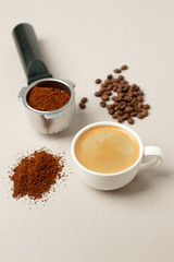 Cup of black coffee next to coffee beans and grains, handle coffee machine filter on gray background, minimal concept, closeup, Morning coffee routine