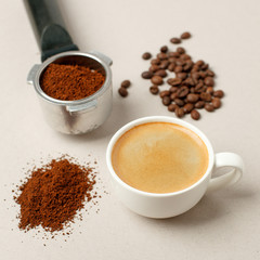 Cup of black coffee next to coffee beans and grains, handle coffee machine filter on gray background, minimal concept, closeup, Morning coffee routine