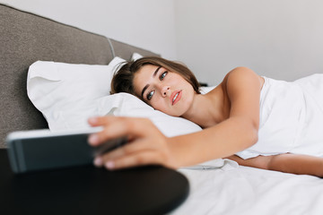 Portrait pretty girl on bed in modern apartment. She looks at phone on table