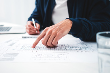 Close up of young architect designer in office working on notebook and building blueprint. Man engineer looking at drawing and computer while sits by the table in loft office