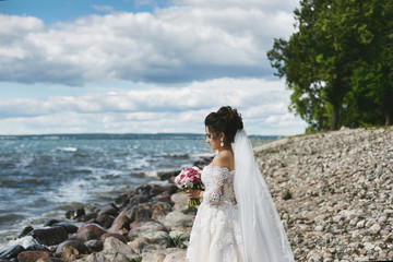 Fototapeta na wymiar Elegant and fashionable brunette plus-size model girl in the stylish lace wedding dress with stylish hairstyle and with the bouquet of flowers in her hands stands at the sea coast