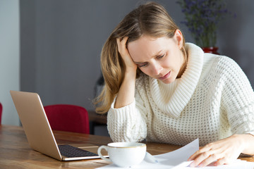 Fototapeta premium Confused young woman reading document at workplace. Upset lady in white sweater analyzing data. Mistake in report concept