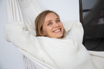 Positive young woman in white sweater resting in home hammock. Happy carefree young woman looking at camera. Vacation concept