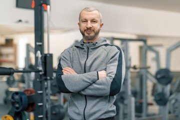Portrait of sporty man with folded hands in gym, handsome bearded trainer looking at the camera