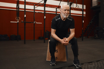 Fototapeta na wymiar Handsome serious male pensioner in sportswear relaxing at fitness center,sitting isolated on wooden box, having confident focused look, preparing for another set of complex muscle building exercises