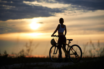 Obraz na płótnie Canvas Back view of woman with bicycle standing on road among grass enjoying the sunset on evening sky. The girl has a beautiful sporty body. Helmet hangs on the handle of bicycle