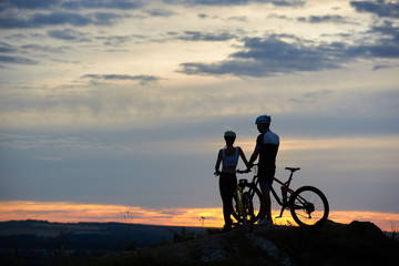 Fototapeta na wymiar Rear view of two young people with mountain bikes stand on top of a cliff with beautiful scenery of hills and sky at sunset. The concept of a healthy lifestyle