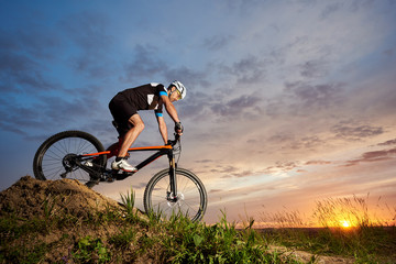 Fototapeta na wymiar Active cyclist wearing sportswear and helmet, riding bicycle alone and rolling down hill. Sporty and robust man cycling against beautiful sunset and rose-blue sky background.