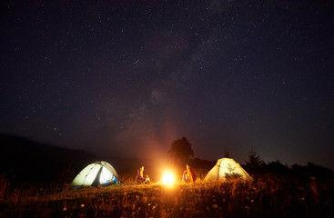 Night camping in mountains. Bright campfire burning between two tourists, boy and girl sitting opposite each other in front of illuminated tents under dark blue starry sky on distant hills background.