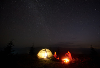 Tourist couple boy and girl sitting on grassy valley in front of brightly lit tent by burning bonfire under starry sky enjoying quiet night in mountains. Tourism, traveling, active lifestyle concept