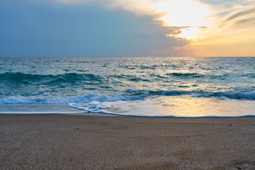 Sunset at the tropical beach, sun behind clouds reflects on water and waves with foam hitting sand.