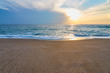 Sunset at the tropical beach, sun behind clouds reflects on water and waves with foam hitting sand.