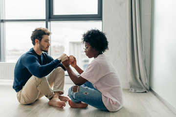 African female yoga instructor giving individual yoga class to young caucasian bearded man, staying indoors, in modern appartment.