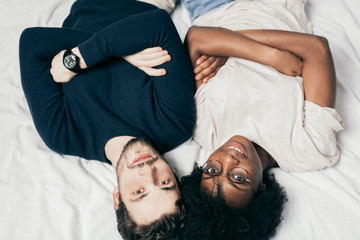 Portrait of happy diverse best friends lying in casual wear with folded hands beside each other on bed and looking at camera. Upside down, top view.