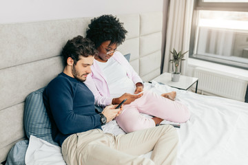 Fototapeta premium Young wife and husband relaxing on bed during their day-off. Spauses of different nationalities sharing funny stories and watching videos using smart phones