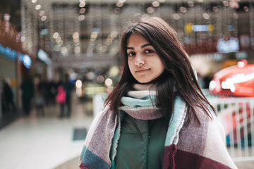 stylish smiley young woman with brown long hair and warm scarf standing in the mall