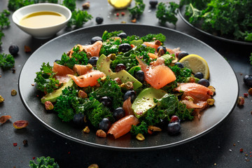 Salmon Kale super food Salad with avocado, pistachio nuts and blueberries on black plate
