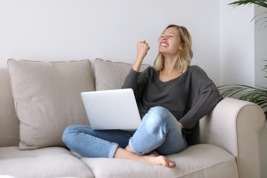 Junge Frau mit Laptop sitzt auf einem Sofa und freut sich