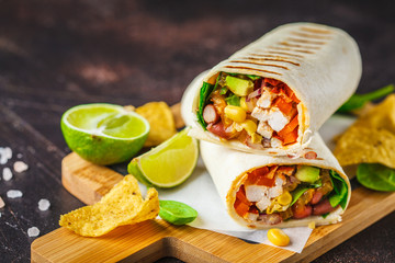 Burritos wraps with chicken, beans, corn, tomatoes and avocado on wooden board, dark background.