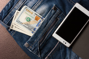 American dollar bills in a jeans pocket and a mobile phone lie on the leather surface.