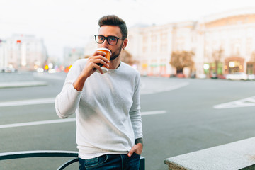 Hipster man standing with takeaway coffee, smiling plesantly, walking on th city street. Happy...