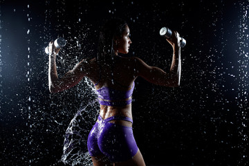 Fototapeta na wymiar Beautiful young girl in purple sportswear poses with dumbbells in aqua studio. Drops of water spread about her fitness body. The perfect figure on the background of water splashes