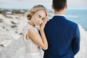 Beautiful young blonde model girl in fashionable white lace dress lean on the handsome man in the stylish blue suit and posing on the white rock at the coast of the Adriatic sea