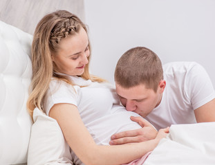 Young husband kisses to the belly of his pregnant wife on the bed