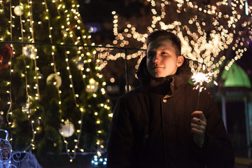 Dreamy handsome man standing on the street with sparkler in hand. Bright bokeh lights of Christmas tree and garlands on background. Winter holidays mood.