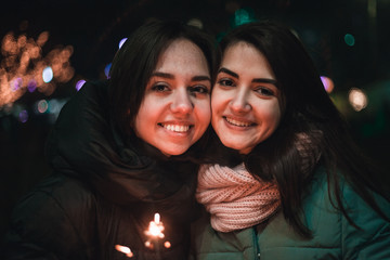 Two positive girlfriends looking at camera on the street with sparkler in hand. Bright bokeh lights of Christmas tree and garlands on background. Winter holidays mood.
