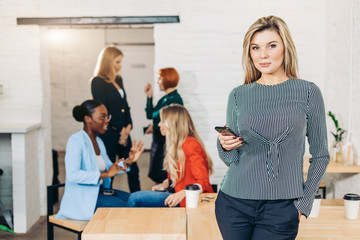 Blonde businesswoman in casual clothes talking on cell phone at modern startup business office interior. Young female group on team meeting on background