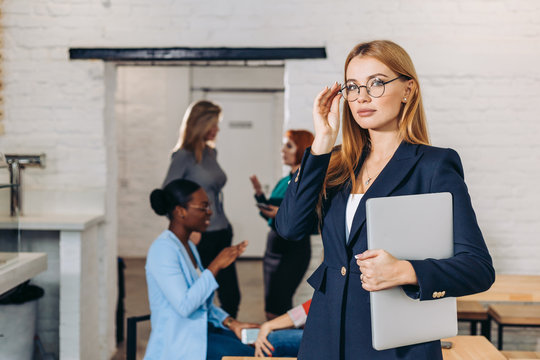 Popular female bloger in spectacles and formal wear holding laptop, thinking about new post, while looking at camera, posing in office with her colleagues on background