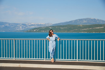 The beautiful girl in a blue dress and sunglasses poses on the bridge, the long chestnut hair, happy and smiles, azure water of the lake and slopes of mountains on a background