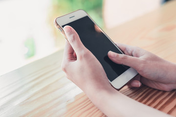 Croppes shot women hand holding white smartphone with blank screen on wooden counter bar in cafe. vintage tone for business technology background concept. closeup mobile.