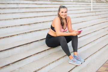 Young smiling plus size woman in sporty top and leggings sitting on stairs with cellphone in hands joyfully looking in camera while spending time outdoor