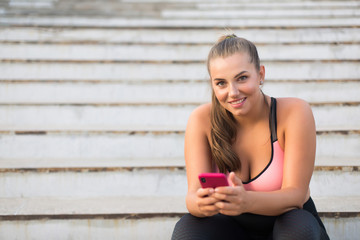 Young pretty plus size woman in sporty top and leggings sitting on stairs with cellphone in hands happily looking in camera while spending time outdoor