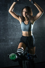 Fototapeta na wymiar Sporty young woman exercising with raised hands while sitting on exercise bike indoors.