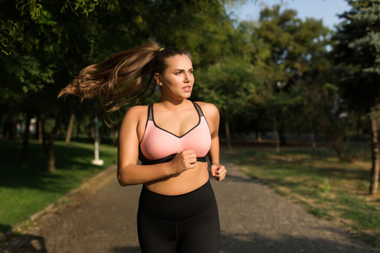 Young attractive plus size woman in sporty top and leggings thoughtfully looking aside while running in cozy city park