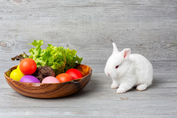 Happy Easter Day. Rabbit with colorful Easter eggs in a wooden tray decorated with vegetables. Cute Easter bunny rabbit with painted Easter eggs on wood background.